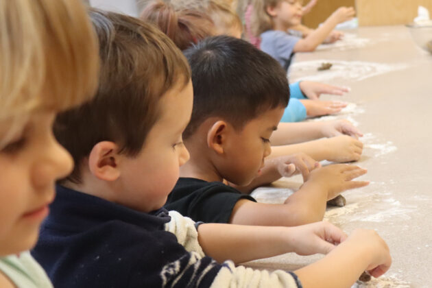 Children sit in a row at a table, using their hands to shape dough on a floured surface.