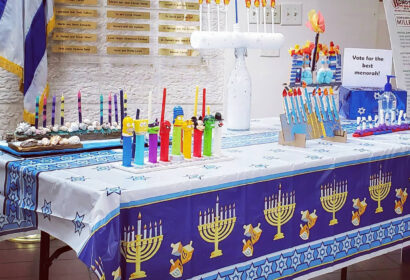 A table decorated for Hanukkah displays menorahs, colorful candles, cookies, and Pez dispensers. A sign invites people to vote for the best menorah. An Israeli flag is visible in the background.