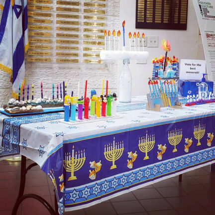 A table decorated for Hanukkah displays menorahs, colorful candles, cookies, and Pez dispensers. A sign invites people to vote for the best menorah. An Israeli flag is visible in the background.