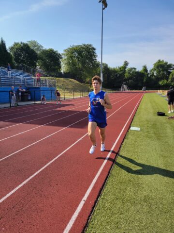 A runner in a blue uniform jogs on a red track at an outdoor stadium on a sunny day, with spectators and other runners in the background.