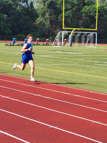 A runner in a blue uniform competes on a red track during a race, with a football goalpost and people in the background on a sunny day.