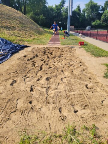 A boy in athletic wear is mid-air above a long jump runway, about to land in a sandpit at an outdoor track and field facility.