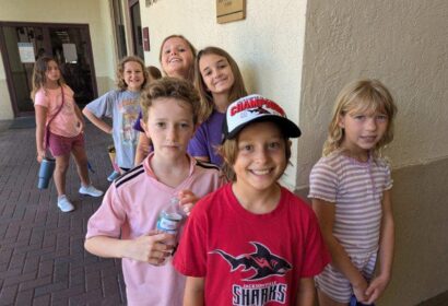 A group of children standing in a line outside a building, wearing casual clothes. One child in front is wearing a red shirt and cap. They appear to be smiling.