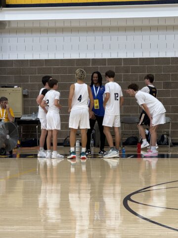 A basketball coach talks to six players in white uniforms during a timeout on an indoor court. Bottles and towels are on the floor nearby.