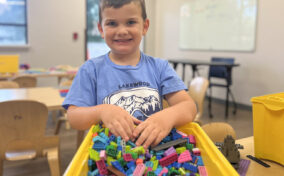 Young boy smiles while standing at a table, his hands in a yellow bin filled with colorful plastic building blocks, in a classroom setting.