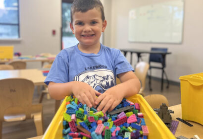Young boy smiles while standing at a table, his hands in a yellow bin filled with colorful plastic building blocks, in a classroom setting.