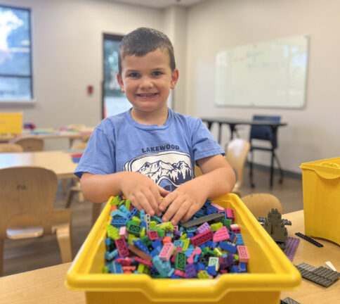 Young boy smiles while standing at a table, his hands in a yellow bin filled with colorful plastic building blocks, in a classroom setting.