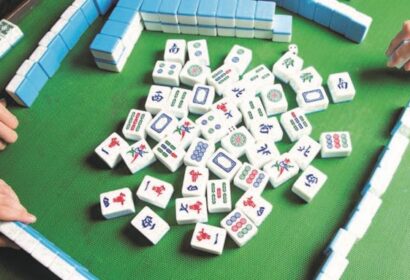 A Mahjong game in progress with three hands visible, tiles arranged on a green playing mat and some scattered in the center.
