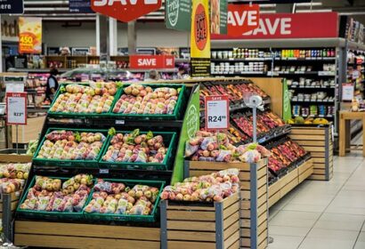 A supermarket produce section displaying various fresh fruits, including apples and pears, arranged in wooden crates. Sale signs and price tags are visible above the produce. Shelves with other products are in the background.