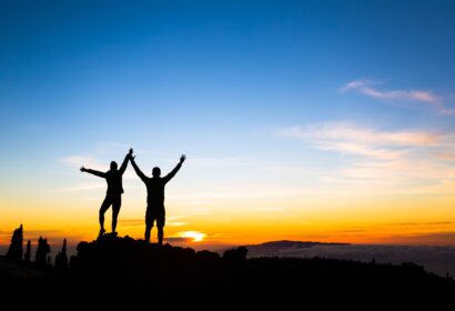 Silhouettes of two people with raised arms stand on a rocky peak at sunrise or sunset, with a colorful sky and distant horizon.