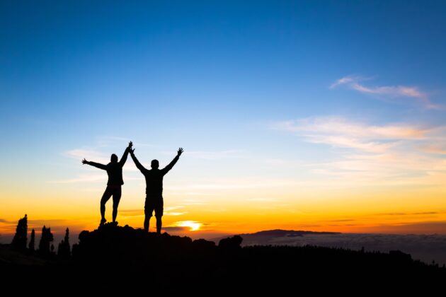 Silhouettes of two people with raised arms stand on a rocky peak at sunrise or sunset, with a colorful sky and distant horizon.