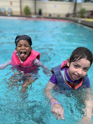 Two children in life jackets stand in a swimming pool. One child is smiling, and the other is playfully sticking out their tongue.