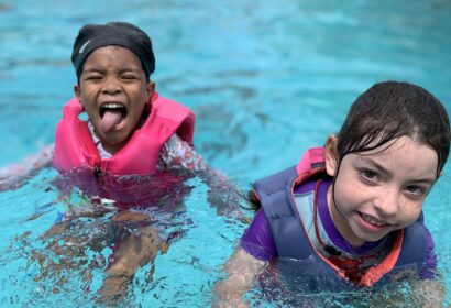 Two children in life jackets stand in a swimming pool. One child is smiling, and the other is playfully sticking out their tongue.