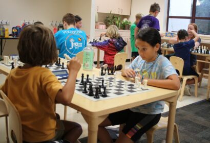 Children playing chess in a classroom setting, seated at tables with chessboards. Volunteers are present, assisting the children.