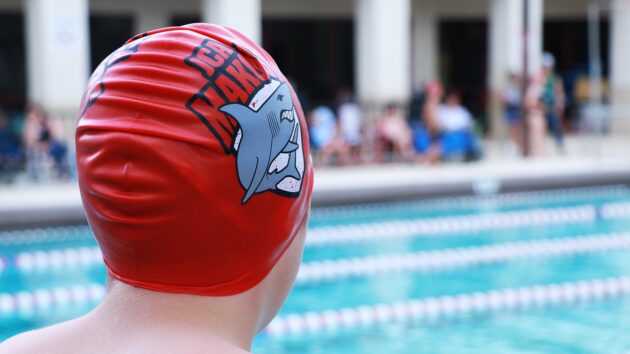 Swimmer wearing a red cap with a shark design looks at an outdoor pool with lane dividers.