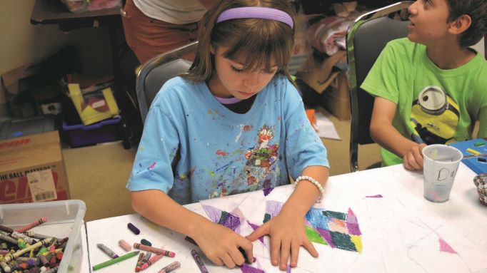Child focuses on coloring a geometric design with crayons at a table, wearing a blue shirt. Another child in a green shirt sits nearby, looking to the side. Art supplies are scattered around at the Jewish Community Alliance.