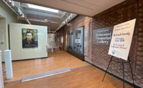 Entrance to the Frisch Family Holocaust Memorial Gallery featuring a brick wall, glass doors, and a sign acknowledging sponsors.