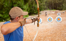 A person in a blue shirt and cap aims a bow and arrow at circular archery targets set up outdoors on a dirt field.