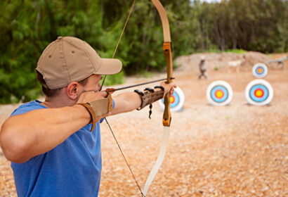 A person in a blue shirt and cap aims a bow and arrow at circular archery targets set up outdoors on a dirt field.