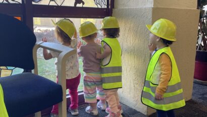 Three toddlers wearing yellow hard hats and safety vests look through a window with stained glass details.