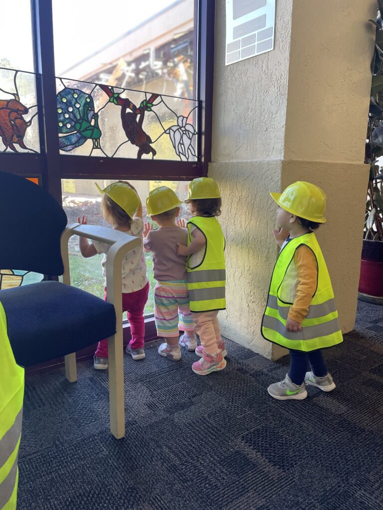 Three toddlers wearing yellow hard hats and safety vests look through a window with stained glass details.