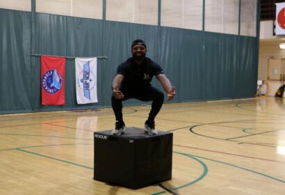 A man squats on top of a black plyometric box in a gymnasium with flags and basketball hoops in the background.