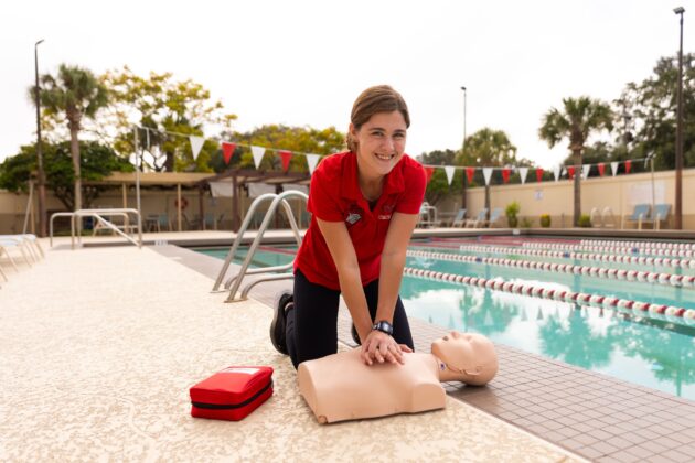 Person demonstrating CPR on a mannequin by a swimming pool, with a red first aid kit nearby.