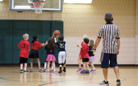 Children playing basketball indoors while a referee watches. A group of young players in red and black shirts compete under the hoop.