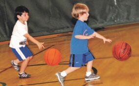 Two young boys dribble basketballs on an indoor court. Both are wearing casual athletic clothing while focusing on their movements.