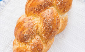 A loaf of braided bread topped with sesame seeds sits on a white textured cloth against a blue wooden background.