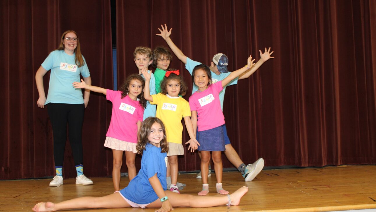A group of children and an adult on a stage posing enthusiastically, with one child doing the splits in front. They wear colorful shirts and have name tags.