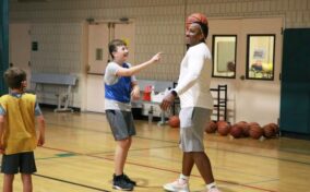 A person holding a basketball on their head interacts with a smiling child in a gym. Other children and basketballs are in the background.