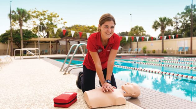 A person practices CPR on a mannequin by a swimming pool. A first aid kit is nearby.
