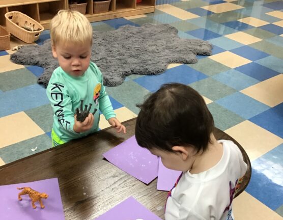 Two toddlers engage with toy animals and purple papers on a wooden table in a classroom with checkered flooring.