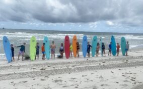 A group of people holding surfboards stands in a line on a sandy beach, with the ocean and a cloudy sky in the background.