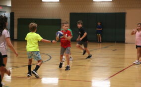 Children playing a game of basketball in a gym, with one child holding the ball while others move around.