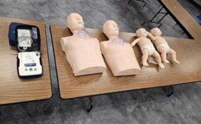 Adult and infant CPR manikins with AED training device on wooden tables in a room with gray flooring.
