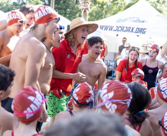 A group of swimmers wearing red caps gather around two people in the center, one in a straw hat. They appear excited and are outdoors with tents in the background.
