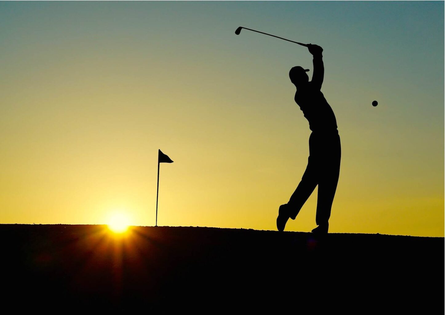 Silhouette of a golfer swinging a club, with a flag in the background and the sun setting on the horizon.