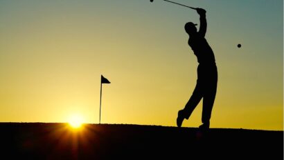 Silhouette of a golfer swinging a club, with a flag in the background and the sun setting on the horizon.