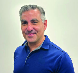 A middle-aged man with short gray hair and a blue collared shirt smiles at the camera against a plain light background.