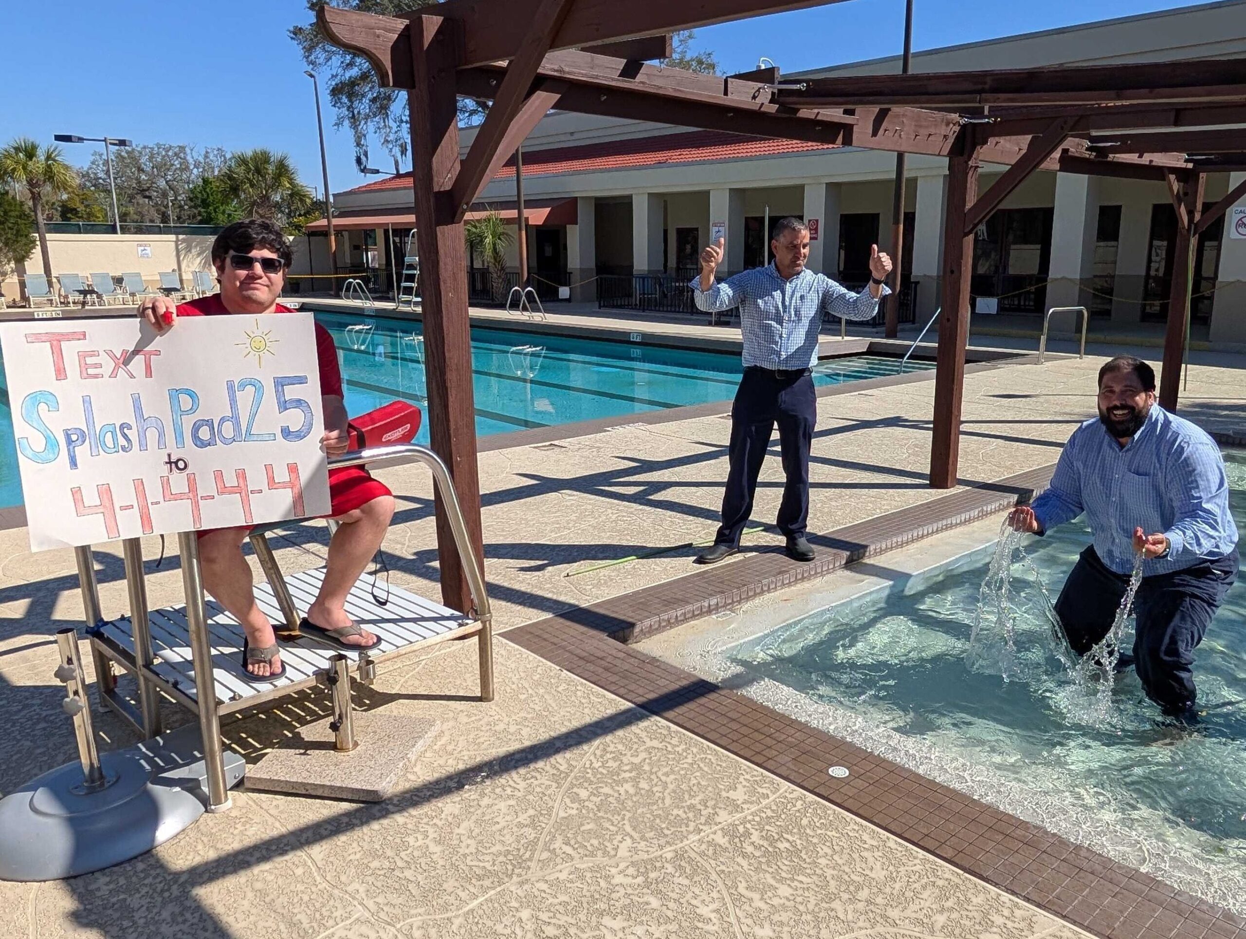 Three men by a pool; one is soaked in a fountain, one stands dry, and a seated man holds a sign with text and numbers. The setting is sunny with buildings in the background.