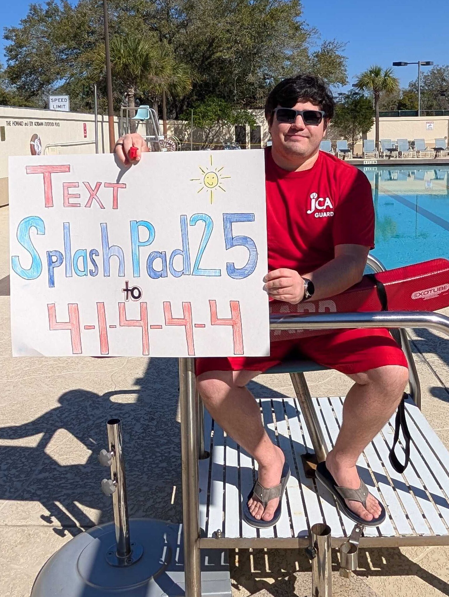 Person in a red lifeguard outfit sits on a lifeguard chair by a pool, holding a sign that says "Text Splash Pad25 to 41444.