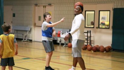 A boy playfully points as a basketball rests on a coach's head in an indoor gym. Another child watches nearby. Basketballs and a chair are in the background.