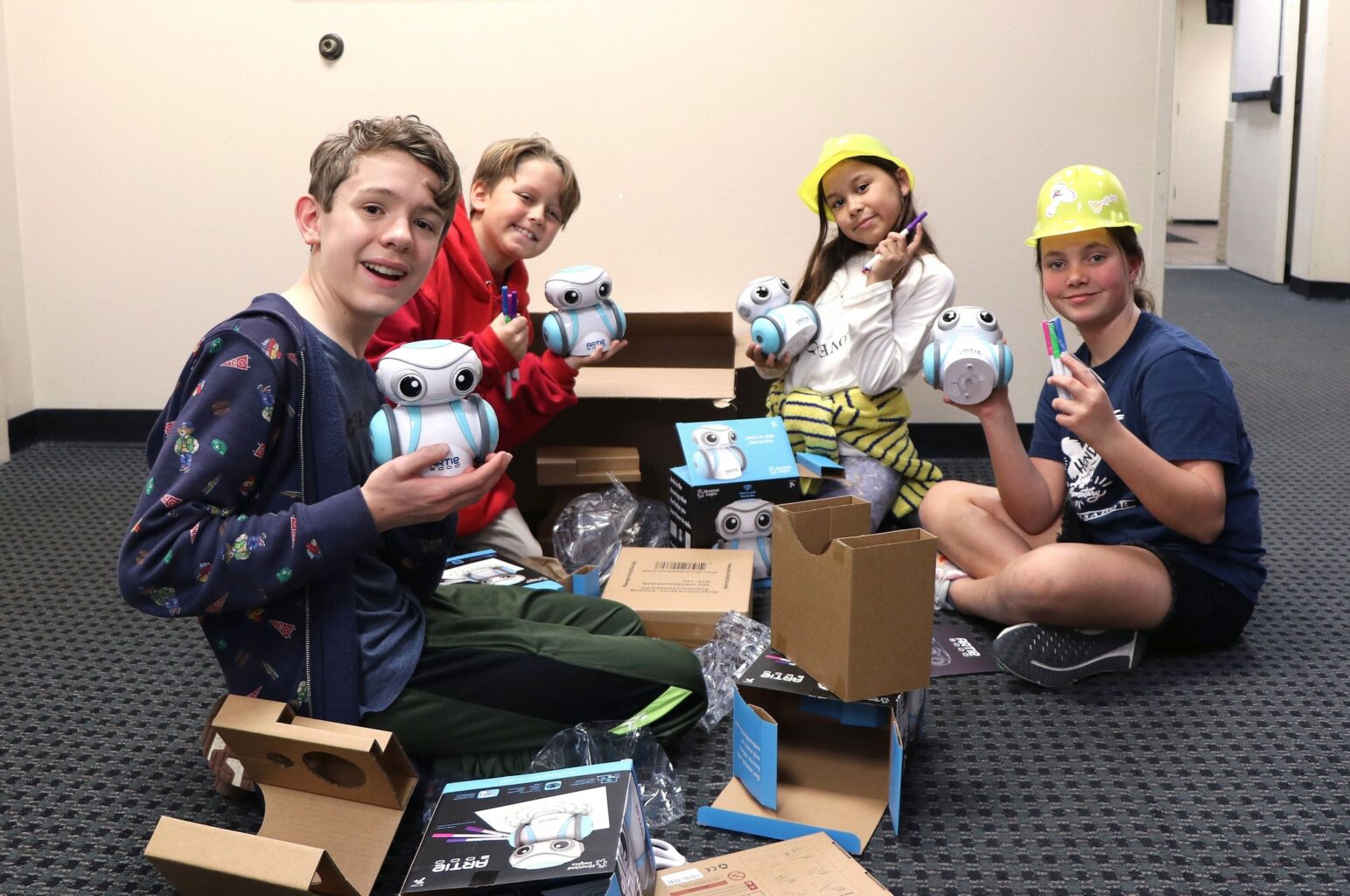 Four children sit on the floor holding round robots and wearing casual clothes. Some are also wearing yellow construction hats. New tech classes will be offered at the JCA in the 2025-26 school year thanks to Johnson and Johnson.