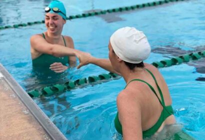 Two swimmers in green swimsuits and swim caps shake hands and smile while standing in a swimming pool near the lane divider.