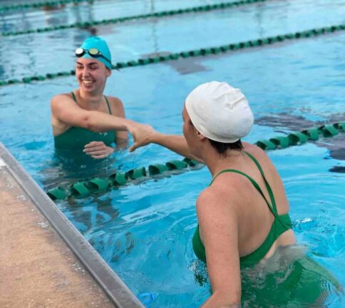Two swimmers in green swimsuits and swim caps shake hands and smile while standing in a swimming pool near the lane divider.