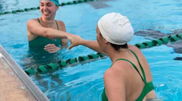 Two swimmers in green swimsuits and swim caps shake hands and smile while standing in a swimming pool near the lane divider.