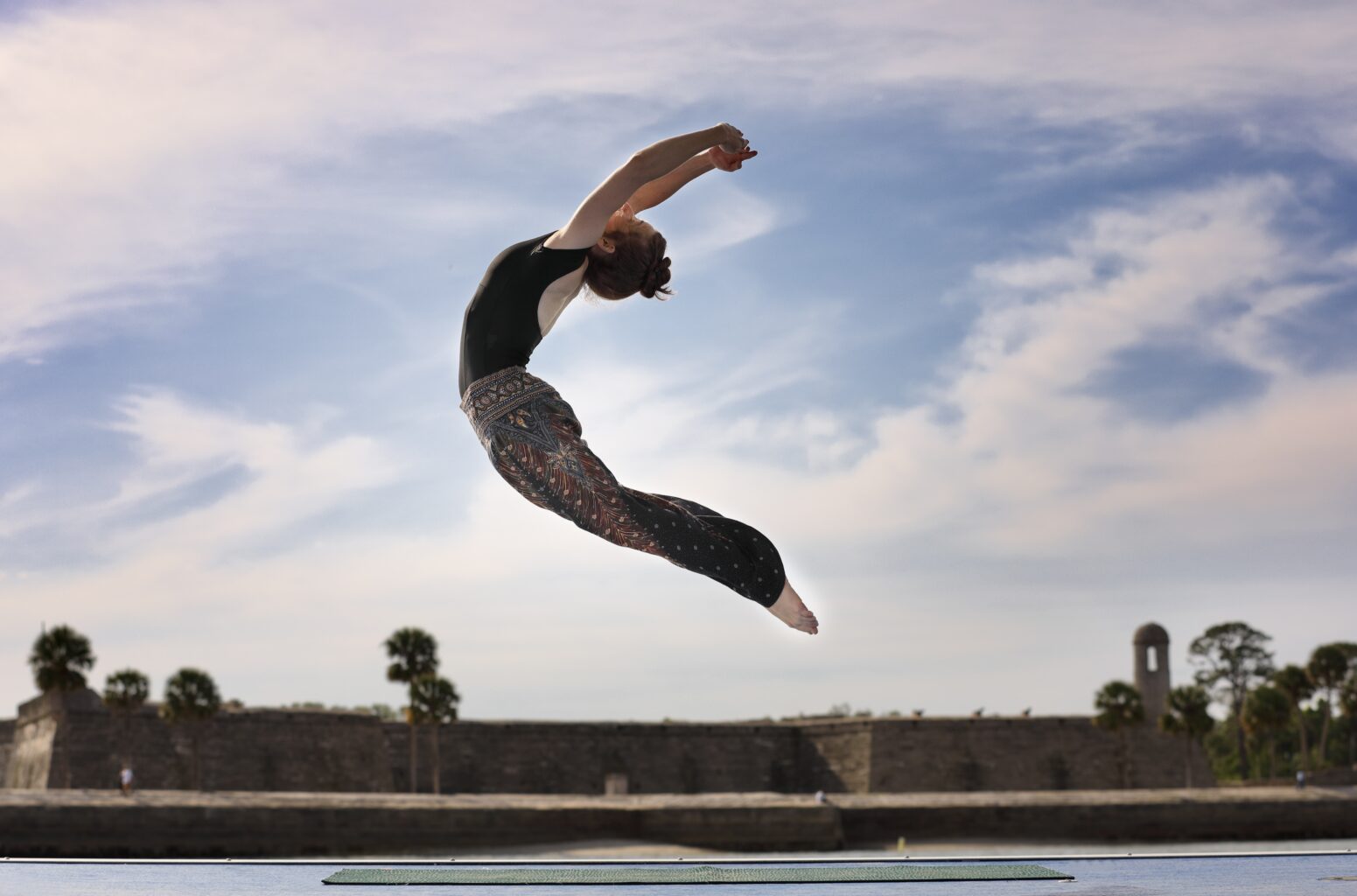 A person performs a mid-air dance or yoga pose outdoors on a mat, with a blue sky and stone wall in the background.