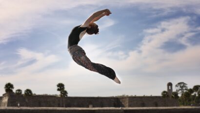 A person performs a mid-air dance or yoga pose outdoors on a mat, with a blue sky and stone wall in the background.